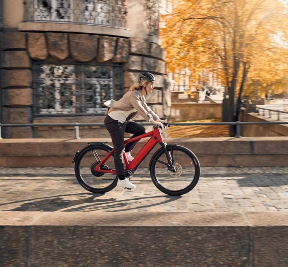Person fährt auf einem roten E-Bike eine Kopfsteinpflasterstraße entlang, neben einem alten Steingebäude und herbstlichen Bäumen.