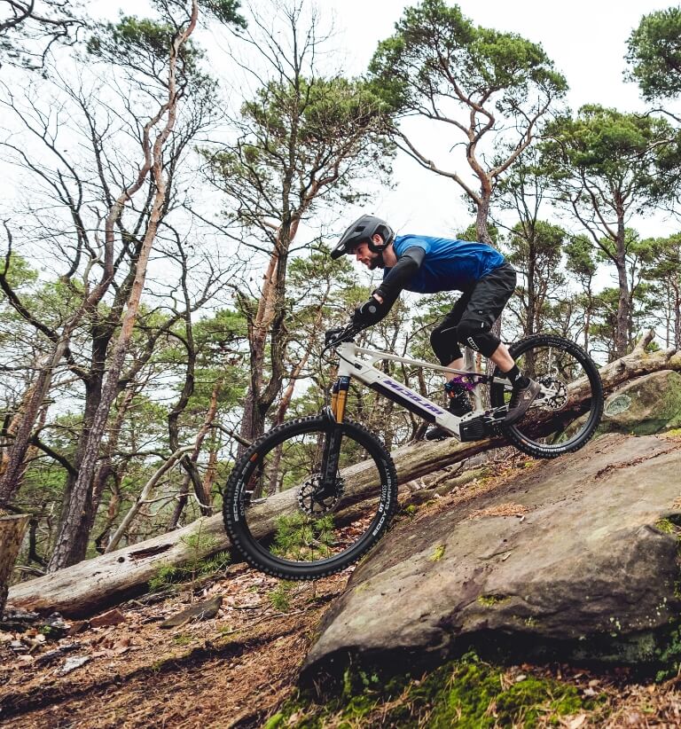 Mountain biker in blue jacket rides down a steep rocky trail in a forest.