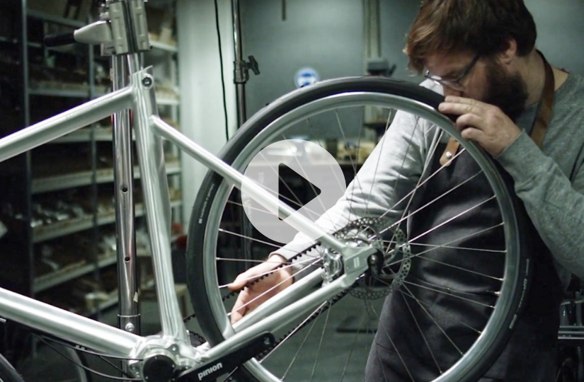 Man inspecting the rear wheel and drivetrain of a silver bicycle in a workshop, with a play button overlay indicating a video.