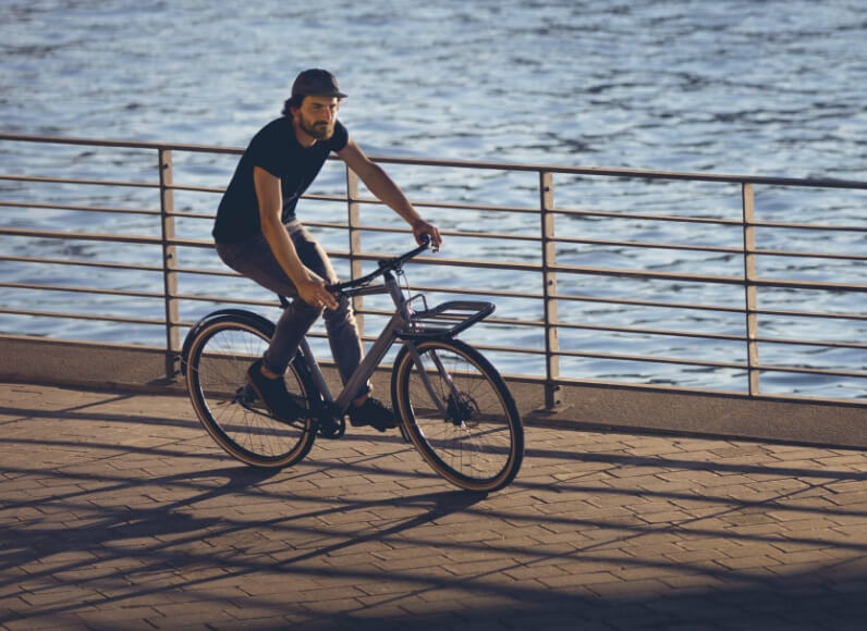 Mann fährt mit dem Fahrrad auf einem Uferweg mit Geländer und Wasser im Hintergrund.