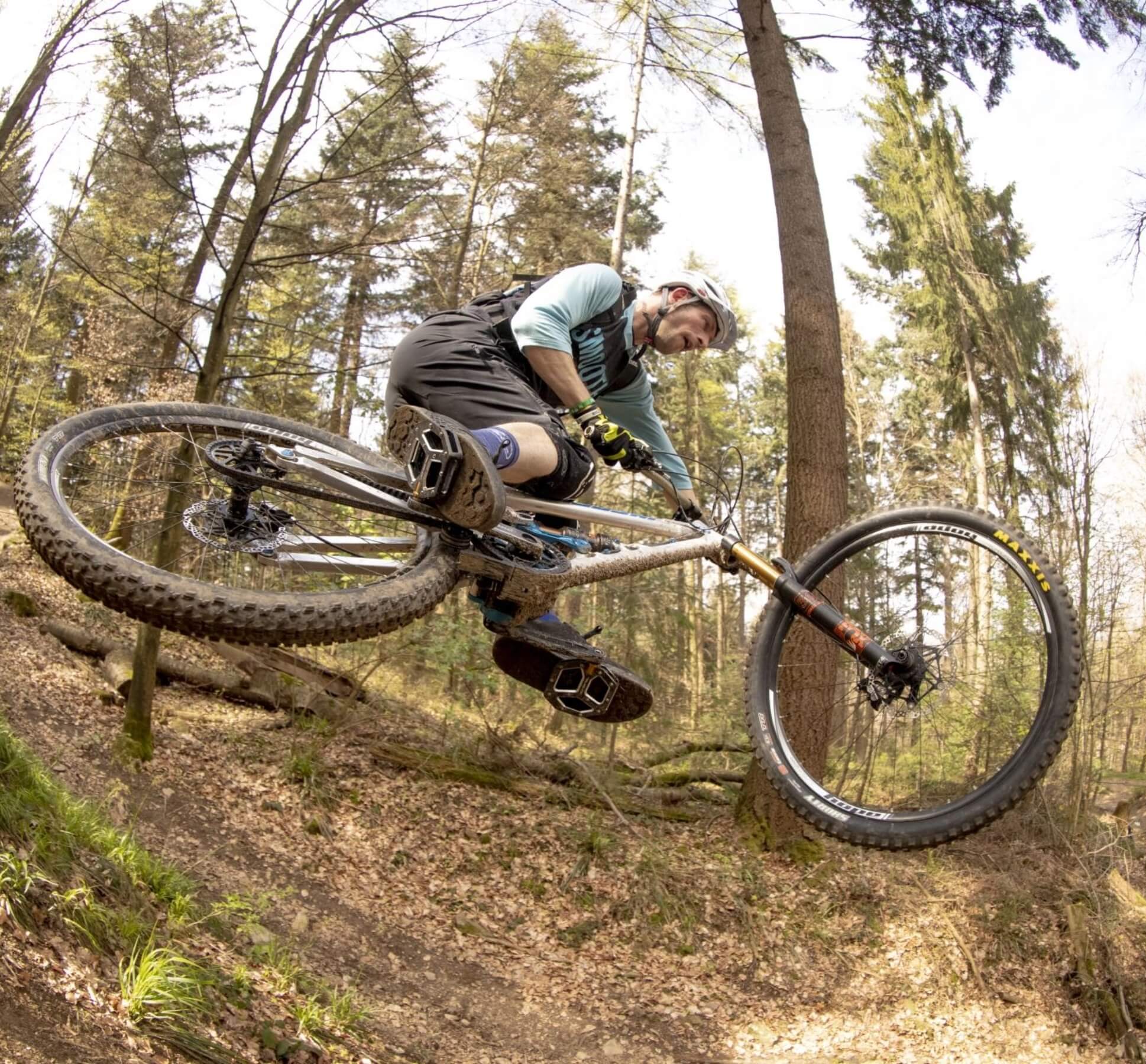 Mountain biker performing a jump on a forest trail, leaning the bike sideways in mid-air.