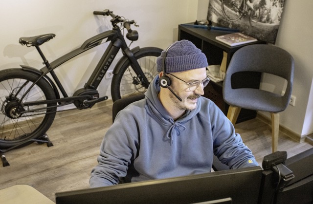 Mann mit Headset und Mütze, der lächelnd am Computer in einem Büro mit einem E-Bike im Hintergrund sitzt.