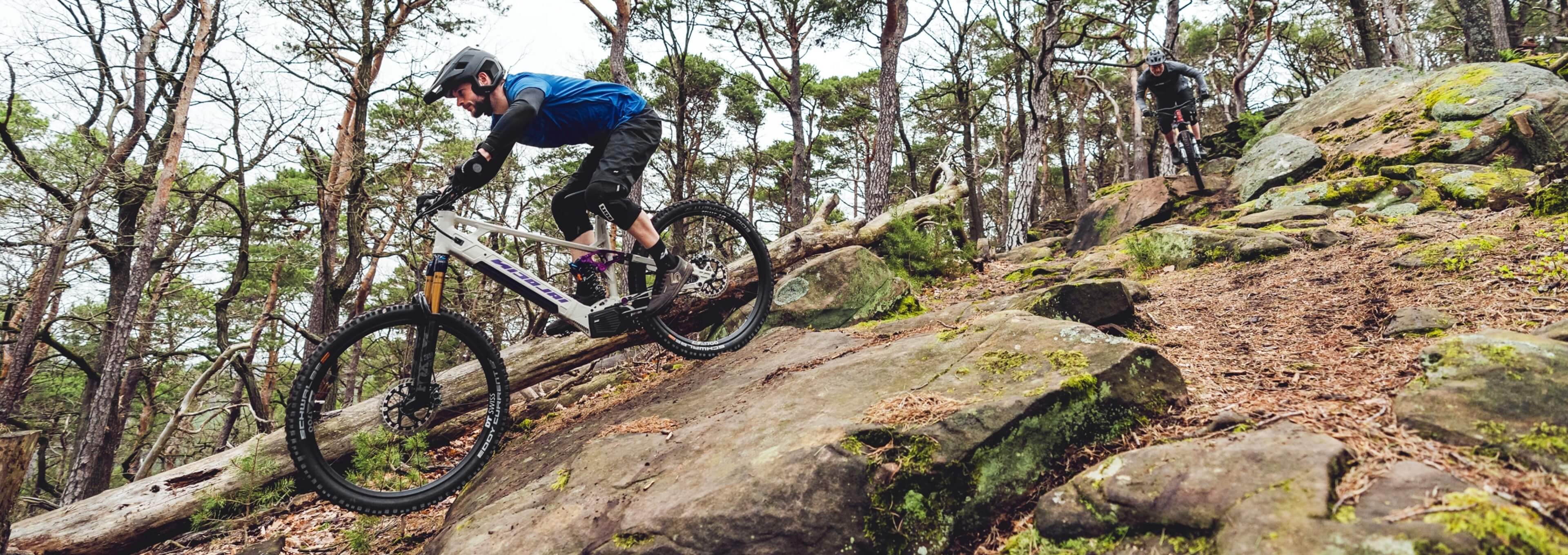 Two mountain bikers ride down a steep rocky trail through a forest.