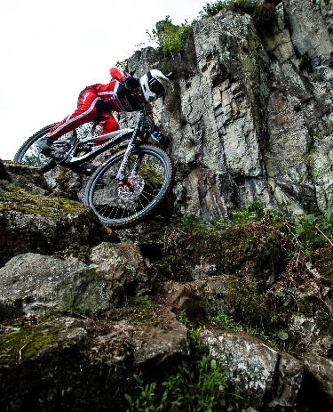 Mountainbiker in roter Kleidung fährt steil einen felsigen Trail an einer Felswand hinab.