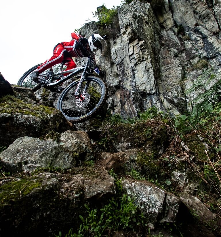 Mountain biker in red gear rides steeply down a rocky cliffside trail.