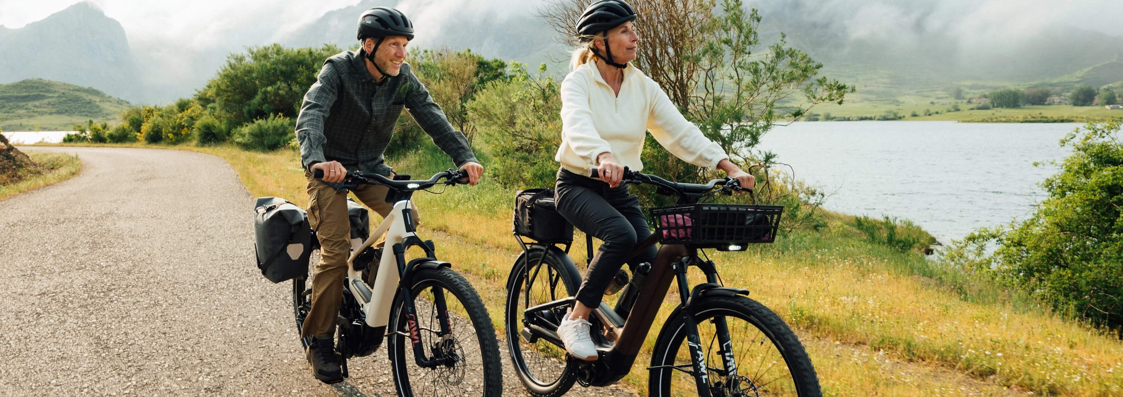 Two people wearing helmets ride electric bikes on a lakeside country road with mountains in the background.