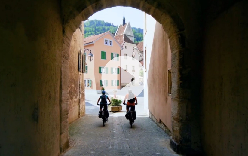Two people riding bikes out of a stone archway toward a town square, with a play button overlay indicating a video.