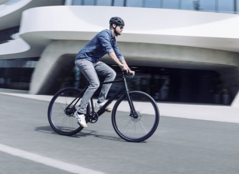 Mann mit Helm fährt mit einem Fahrrad auf einer Straße vor einem modernen Gebäude.